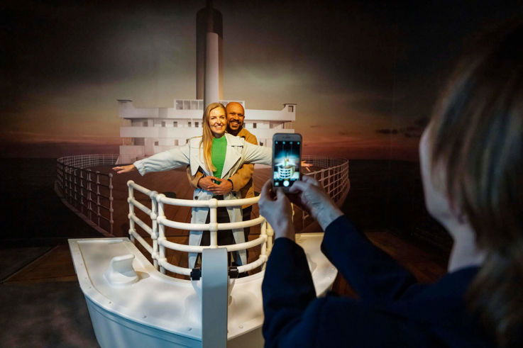A woman photographing a couple posing on a boat, capturing a moment on the water at the Titanic Belfast experience.